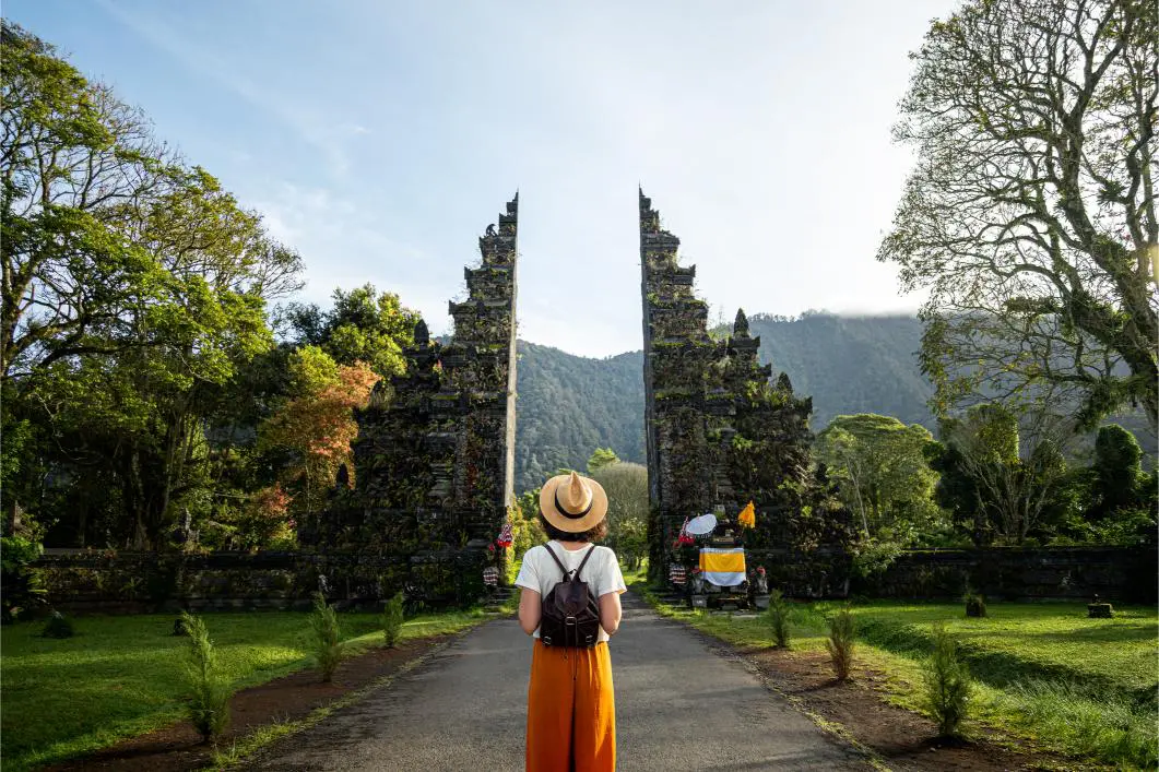 Person wearing a hat and backpack stands in front of a traditional stone gate surrounded by lush greenery.