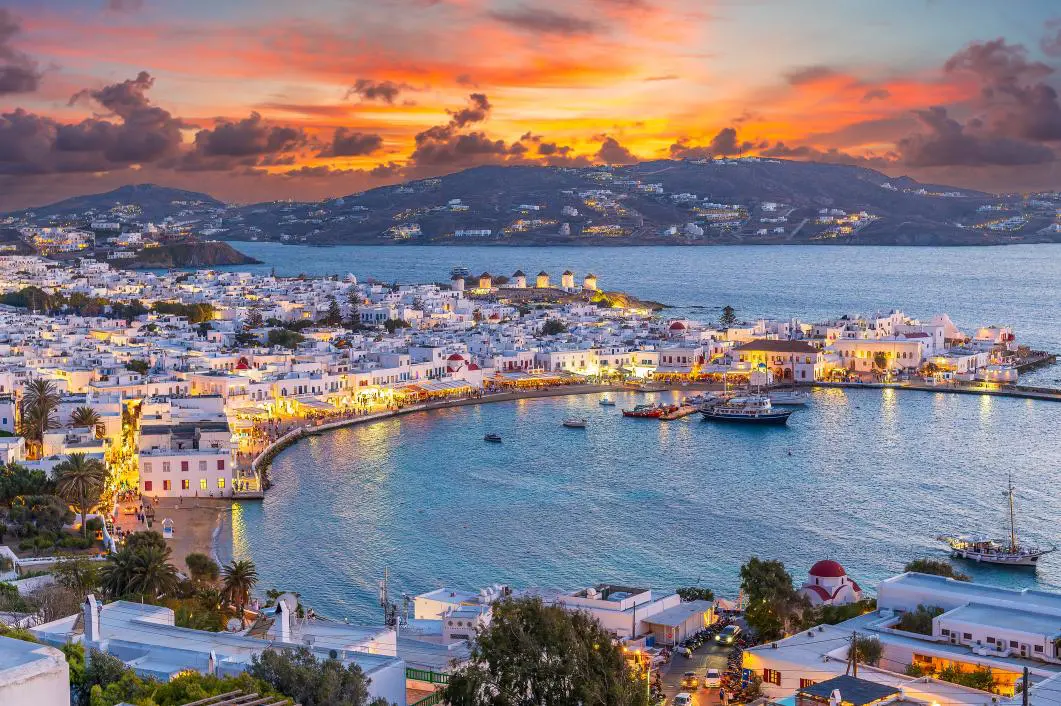 Sunset view of a coastal town with white buildings and a harbor, surrounded by hills and calm water.