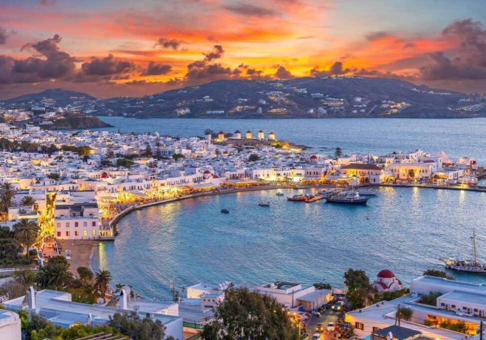 Sunset view of a coastal town with white buildings and a harbor, surrounded by hills and calm water.