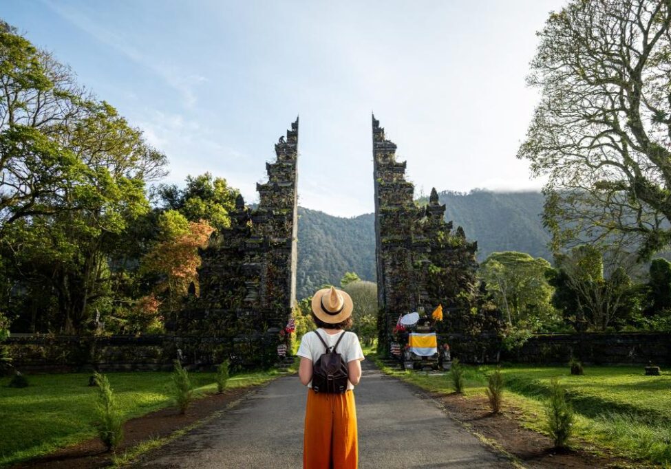 Person wearing a hat and backpack stands in front of a traditional stone gate surrounded by lush greenery.